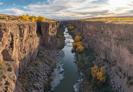A scenic view of a narrow canyon with a flowing river, flanked by steep rocky walls and yellow autumn trees under a blue sky. - Powered by Adobe