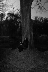 Woman in a black dress sitting by a tree in a serene outdoor setting.