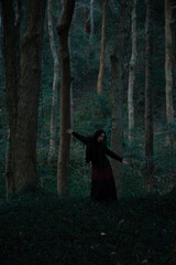 Woman in a dark forest wearing a black long dress, standing with arms outstretched, conveying a sense of serenity.