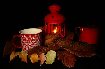 Autumn still life with red cup, lantern and candle among dry leaves on dark background. Warm seasonal mood evoking fall evenings and cozy atmosphere, also suitable for Halloween themes.