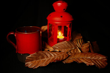 Autumn still life with red cup, lantern and candle among dry leaves on dark background. Warm...