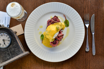 veal souvlaki with poached egg in a white plate on a table with cappuccino and a retro clock