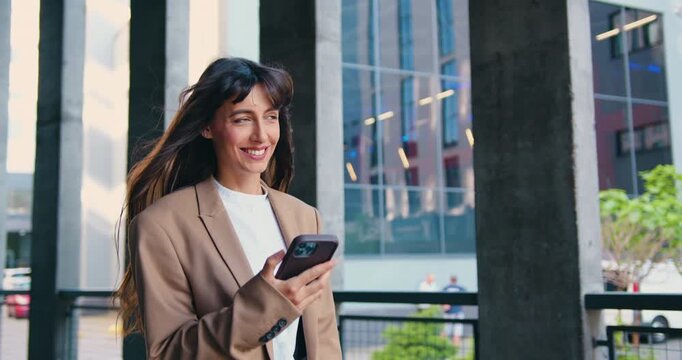 Confident and smiling young businesswoman walking outside modern office building, smiling while using smartphone, checking messages, browsing apps, staying connected on the go, outdoor