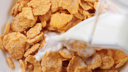 Close-up of crispy cornflakes with milk being poured over them in a bowl. Breakfast food concept with fresh cereal, dairy and morning meal preparation. Healthy start to the day. Textured background