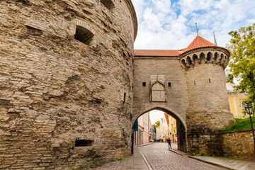 Medieval stone city gate with tower and cobblestone street in historic European old town under partly blue sky, perfect for travel, architecture, and history themes