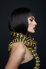 In a dramatic dark setting, a woman with a sleek black bob hairstyle poses confidently. She has bold makeup, and a yellow and black striped snake rests around her neck, creating a striking contrast