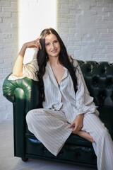 woman with long dark hair sits comfortably on a green couch. She wears casual striped pajamas and poses thoughtfully as soft light illuminates the room