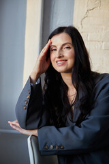 woman with long black hair smiles while resting her hand on her forehead. She wears a stylish gray blazer and sits comfortably in an elegant indoor location, exuding confidence and charm