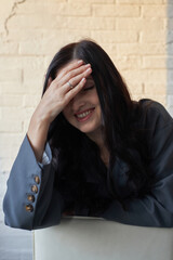woman with long black hair smiles while resting her forehead on her hand. She sits casually in a bright, modern indoor space, looking relaxed and happy