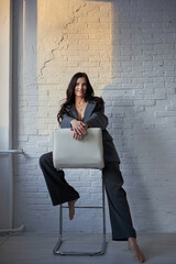 woman in a stylish suit sits on a stool, one leg draped over it, smiling at the camera. Soft sunlight highlights her features against the plain brick backdrop