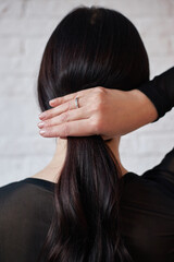 woman is seen with her back to the viewer, gently holding her long dark hair with one hand while wearing a black top. background features a light-colored wall