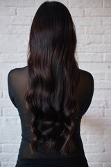 person stands with their back facing the viewer, showcasing long wavy dark hair cascading down. They wear a black top, and the backdrop is a clean white brick wall