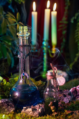 Two potion bottles sit on mossy ground, emitting wisps of smoke. Surrounding them are green plants, lit candles in the background, and a hint of mystery in the air