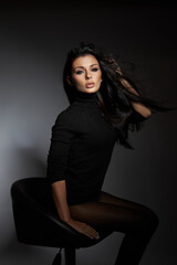 In a dimly lit studio, a model with long black hair strikes a confident pose on a chair, dressed in a sleek black top. atmosphere is elegant and artistic