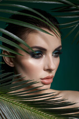 woman showcases her striking makeup and smooth skin while partially hidden behind lush green palm leaves. scene emphasizes a vibrant, tropical vibe and beauty aesthetics