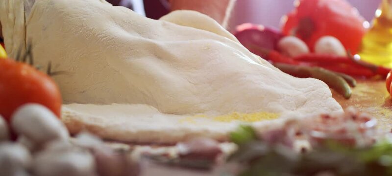 Professional baker's hands kneading and stretching fresh dough for an authentic italian pizza on a wooden table surrounded by fresh ingredients like tomatoes, mushrooms, and olive oil