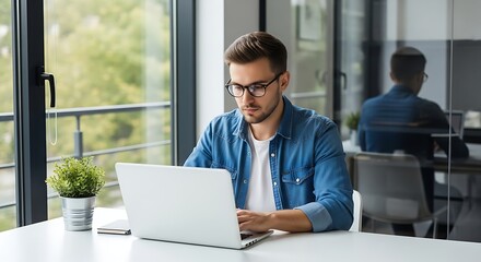 Fototapeta premium Young professional working on laptop in modern office space with greenery outside