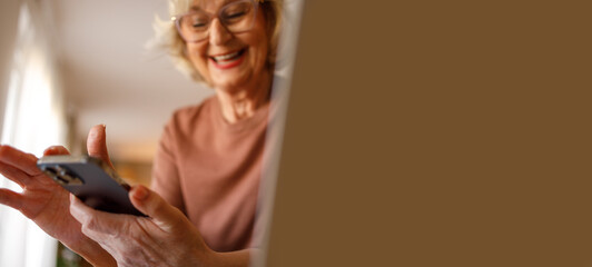 Low angle view of cheerful senior woman checking text messages over smart phone while sitting at home