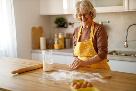 Cheerful elderly woman sprinkling flour on wooden counter and preparing dough for traditional cooking at home