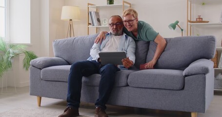 Smiling seniors cuddle on a gray sofa at home, browsing a tablet, sharing jokes, and enjoying technology and online connection in retirement. Senior couple browsing internet together.