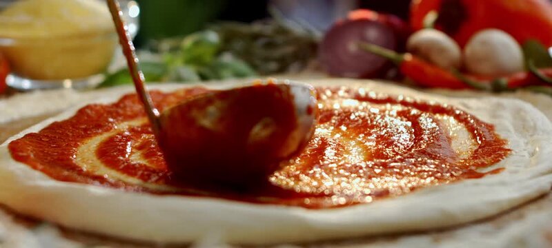 Vertical video of close up of a professional chef spreading fresh tomato sauce with a ladle on a raw pizza dough, the first step in preparing a delicious homemade traditional italian margherita