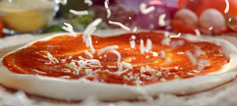 Grated mozzarella cheese slowly falling onto a raw pizza base covered with fresh tomato sauce. Close-up slow-motion shot of the pizza making process with fresh ingredients in the background
