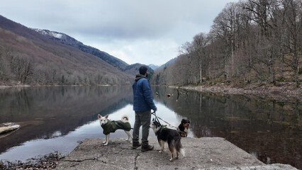 A European man standing with his back to the camera, accompanied by three dogs, gazing into the distance at a calm autumn lake.