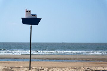 Wooden model of boat on pole on sandy beach with real big cargo ship in background, sunny day