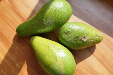 Three green avocados on a wooden surface with sunlight casting shadows