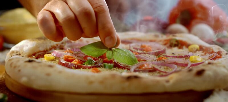 Vertical video of professional chef's hand adding fresh basil leaves to a steaming hot italian pizza with salami, red onions, and vegetables, ready to be served in a restaurant or pizzeria
