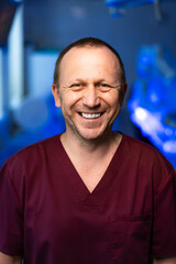 Smiling male doctor in scrubs standing in hospital room. Portrait of a happy male doctor in burgundy scrubs smiling in a hospital.