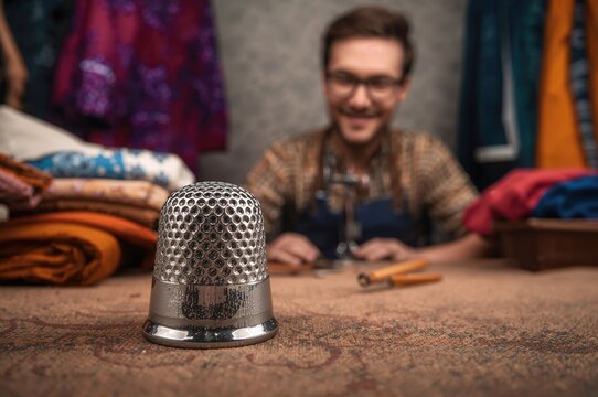 Close-up of silver sewing thimble on a textured surface with colorful fabric piles in the background and a tailor smiling while working on clothing design and sewing project tools - Powered by Adobe