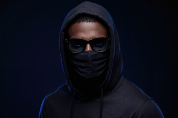man wearing dark hoodie, black sunglasses, and face mask against a dark blue illuminated background highlighting urban fashion and anonymity