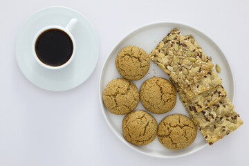 A white plate with oatmeal cookies and seed crackers served next to a cup of black coffee on a white background. Minimalist breakfast or snack concept.