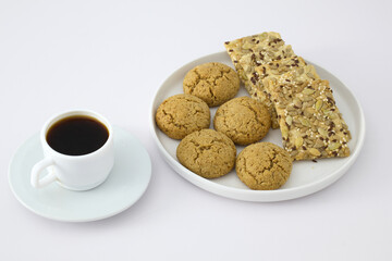 A white plate with oatmeal cookies and seed crackers served next to a cup of black coffee on a white background. Minimalist breakfast or snack concept.