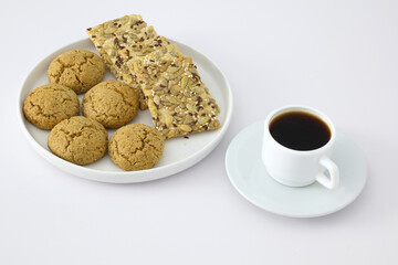 A white plate with oatmeal cookies and seed crackers served next to a cup of black coffee on a white background. Minimalist breakfast or snack concept.