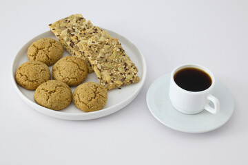 A white plate with oatmeal cookies and seed crackers served next to a cup of black coffee on a white background. Minimalist breakfast or snack concept.