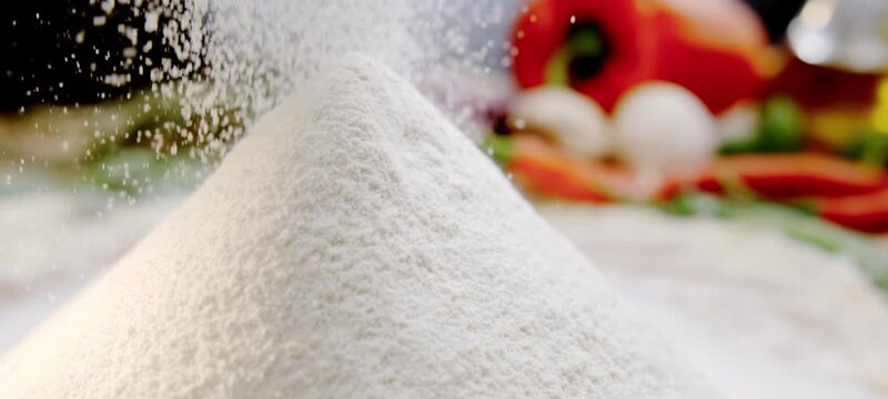 Close-up shot of fine white flour being sieved onto a pile on a kitchen counter, with fresh vegetables blurred in the background, representing the process of preparing dough for baking