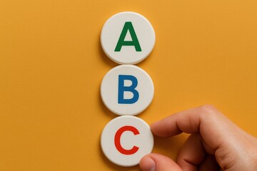 A human hand gently positions the letter "C" disc, completing a stack of "A", "B", and "C" alphabet letters on a vivid yellow surface, creating a learning concept.