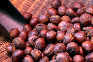 Close-up of roasted chestnuts with cracked, glossy shells in a wicker basket. A metal scoop in the corner hints at serving, with warm lighting creating a cozy, autumn street food vibe.