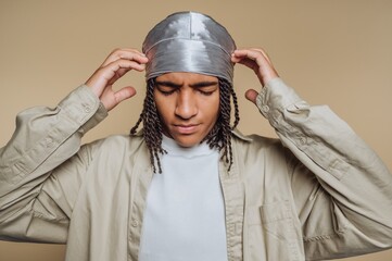 Young man wearing a silver durag and looking thoughtful while standing against a neutral background