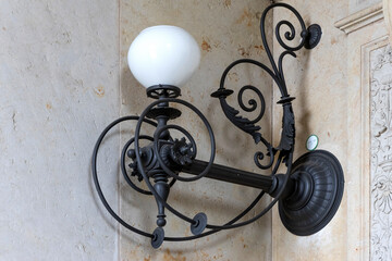 Bildüberschrift:
Ornamental wrought iron wall lamp with white glass shade mounted on stone wal