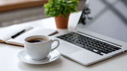 Warm coffee beside a laptop on a desk with a plant and a notebook in a cozy workspace setting