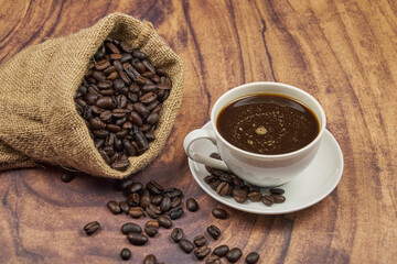 Coffee cup, foreground, with freshly roasted beans in a jute sack on a rustic wooden table with copy space