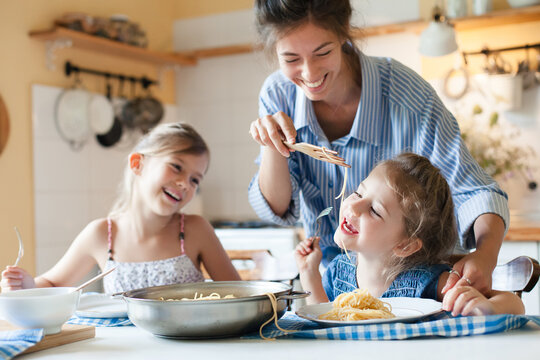 Happy mother and kids eating pasta at home kitchen, having fun, enjoying family lunch, healthy comfort food for children. Homemade Italian cuisine, mac and cheese. Candid, authentic lifestyle moment