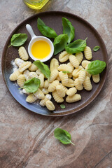 Plate with uncooked potato gnocchi and fresh green basil, vertical shot on a brown granite background, top view