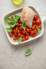 Roasted cherry tomatoes with fresh bread and green basil on a beige stone background, vertical shot, above view