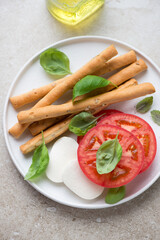 White plate with grissini breadsticks and ingredients for caprese, vertical shot on a beige stone background, middle close-up