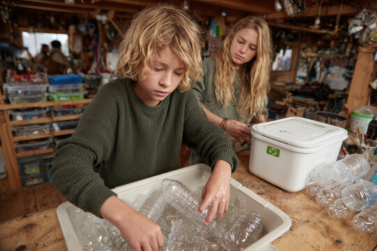 A young boy rinsing a plastic bottle at the sink while his older sister places clean recyclables into a modern recycling station, warm natural light, candid eco-friendly routine - Powered by Adobe