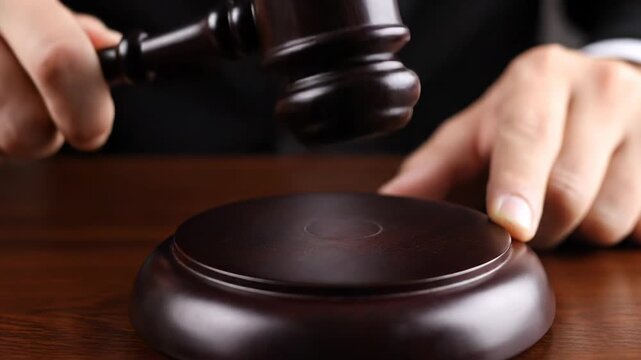 Close up shot of a judge's hands striking a wooden gavel on a sounding block. The decisive tap signals the end of a court proceeding as judgment is passed